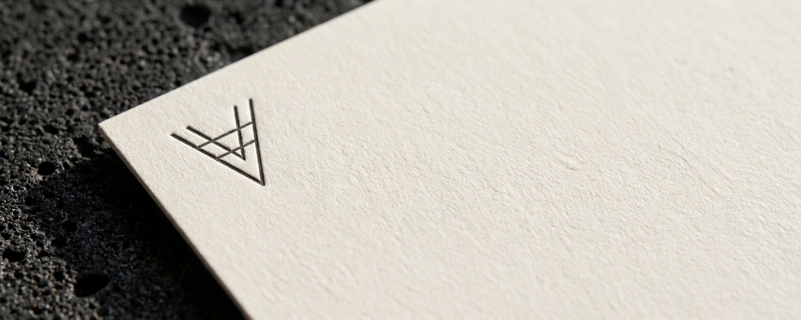 Extreme close-up of blind-debossed V monogram on 350gsm cotton card stock under raking daylight